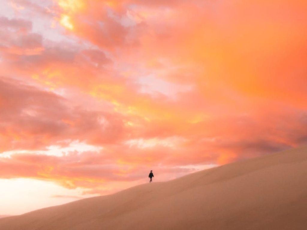 unrecognizable tourist walking along hilly desert under sunset sky